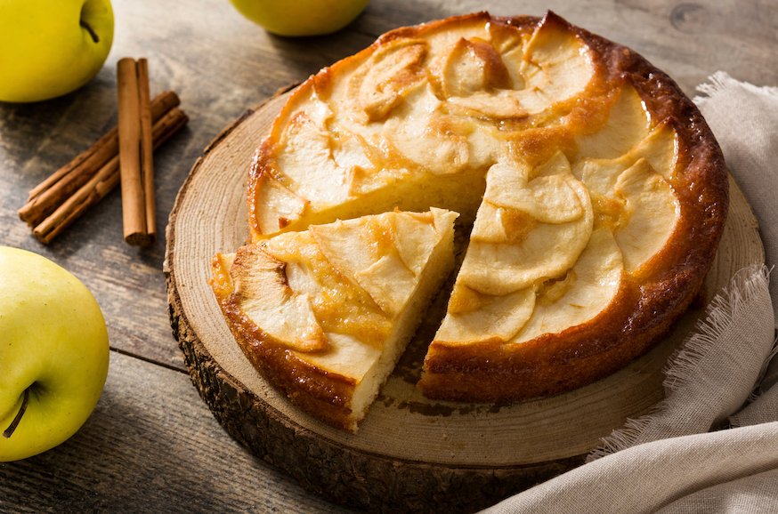 Gâteau moelleux aux pommes et à la frangipane, doré et fondant, présenté sur une planche en bois. Des bâtons de cannelle et des pommes jaunes l'entourent, sur une table rustique en bois.